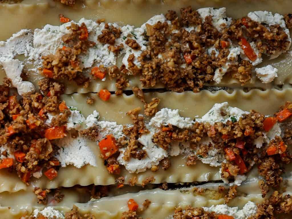 Close-up of uncooked lasagna layers with dollops of ricotta cheese, ground vegan meat, and chopped red carrots. The ingredients are spread unevenly on the pasta sheets, showcasing the textures and colors of the dish.